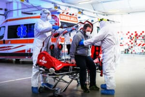 Due operatori sanitari con tutta integrale, mascherina e occhiali protettivi, visitano un paziente durante il covid seduto su una barella con alle spalle l'ambulanza. 